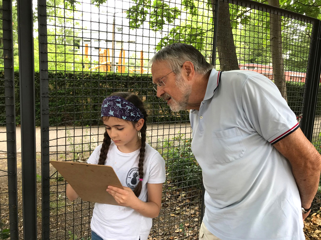 Et barn holder et clipboard og kigger på det, mens en ældre person læner sig ind for at se på clipboardet. De står udendørs nær et metalhegn med træer og grønt i baggrunden.