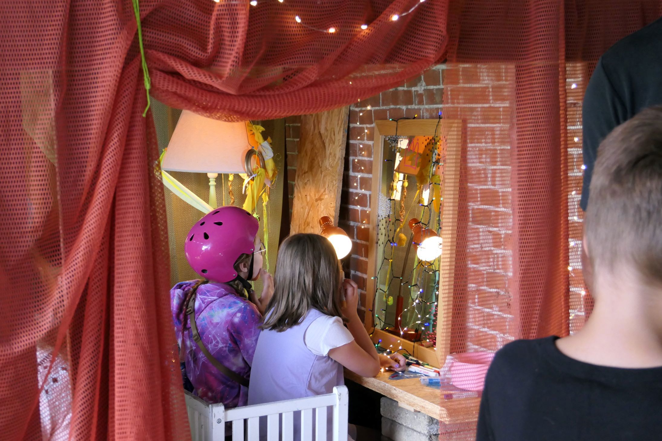 To børn sidder ved et lille bord med et spejl, omgivet af røde gardiner og lyskæder. Et barn har en pink hjelm på, og det andet lægger makeup. 