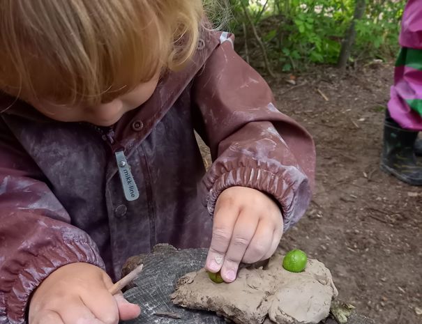 Et barn i en brun jakke leger med ler på en træstub i naturen. Barnet placerer en lille grøn genstand i leret.