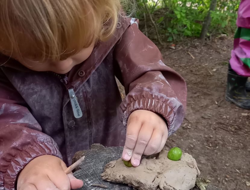 Et barn i en brun jakke leger med ler på en træstub i naturen. Barnet placerer en lille grøn genstand i leret.