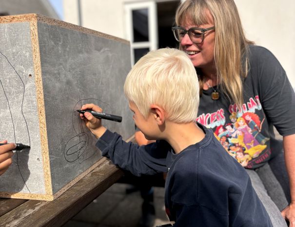 Et barn tegner på en træoverflade med en sort tusch, mens en voksen står i nærheden og observerer. I baggrunden ses en del af en bygning med hvide vægge og rødt tegltag.