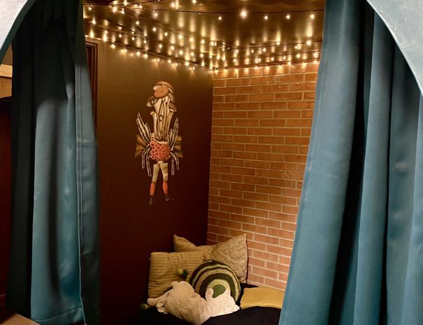 En hyggekrog med lyskæder hængende fra loftet, der skaber en varm og indbydende atmosfære. Væggene er dekoreret med murstens tapet, og der er en collage af en person lavet af svampe på en væg. 