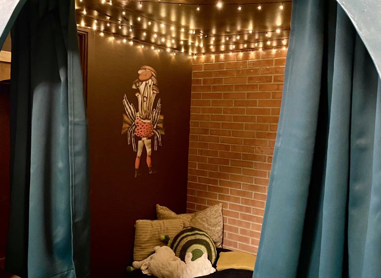 En hyggekrog med lyskæder hængende fra loftet, der skaber en varm og indbydende atmosfære. Væggene er dekoreret med murstens tapet, og der er en collage af en person lavet af svampe på en væg. 