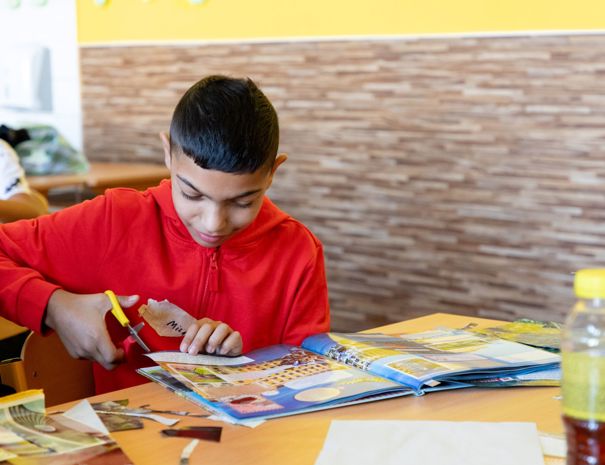 Et barn i en rød hættetrøje sidder ved et bord og klipper papir med en saks. Der er bøger og papirer spredt ud på bordet, og en anden person er delvist synlig til højre i billedet. Baggrunden inkluderer en gul væg og træpaneler.