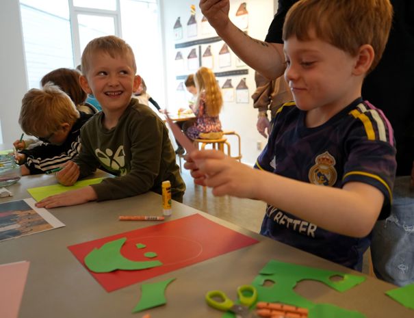 En gruppe børn, der laver kunsthåndværk ved et bord med farvet papir, sakse, limstifter og farveblyanter. En voksen hjælper børnene, mens de klipper former ud af grønt papir og tegner eller samler deres kunstværker.