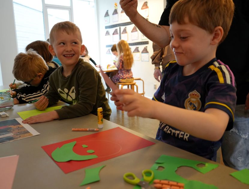 En gruppe børn, der laver kunsthåndværk ved et bord med farvet papir, sakse, limstifter og farveblyanter. En voksen hjælper børnene, mens de klipper former ud af grønt papir og tegner eller samler deres kunstværker.