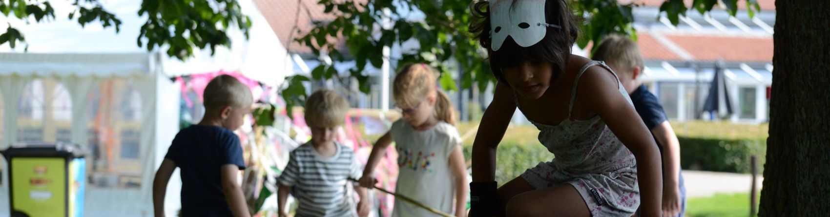 En gruppe børn leger udendørs i en park. Et barn bærer en maske og klatrer på en stor sort struktur, mens andre børn leger i baggrunden.