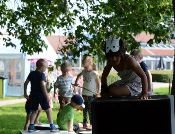 En gruppe børn leger udendørs i en park. Et barn bærer en maske og klatrer på en stor sort struktur, mens andre børn leger i baggrunden.