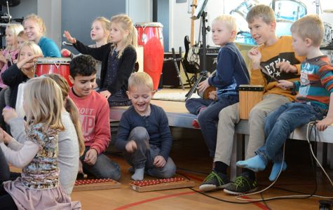 Børn sidder på gulvet og på en scene og spiller på musikinstrumenter. Baggrunden inkluderer flere musikinstrumenter.