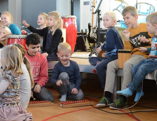 Børn sidder på gulvet og på en scene og spiller på musikinstrumenter. Baggrunden inkluderer flere musikinstrumenter.
