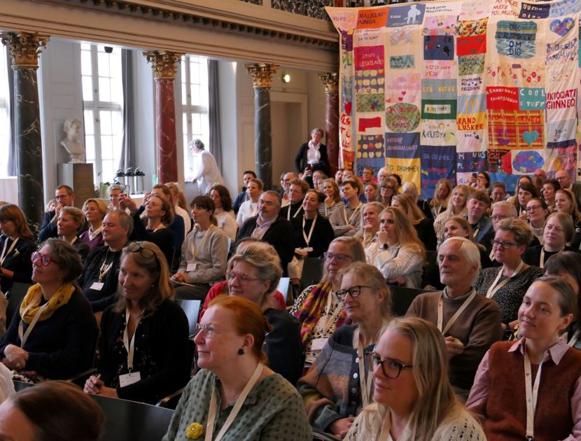 En stor gruppe mennesker sidder i et auditorium eller konferencerum med udsmykkede søjler og højt til loftet. Der hænger et farverigt kunstværk af sammensyede bannere på væggen til højre.