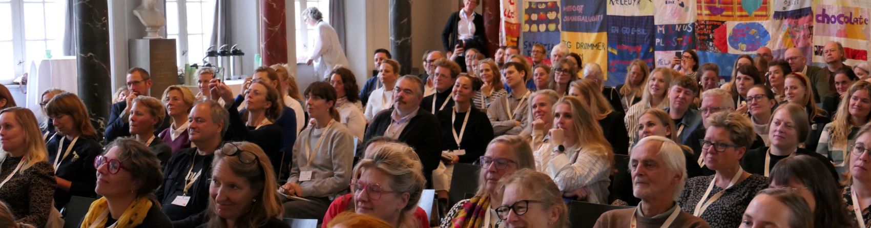 En stor gruppe mennesker sidder i et auditorium eller konferencerum med udsmykkede søjler og højt til loftet. Der hænger et farverigt kunstværk af sammensyede bannere på væggen til højre.