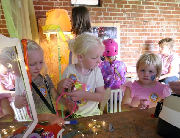 En gruppe børn er engageret i en aktivitet ved et bord. Indendørs omgivelser med en murstensvæg i baggrunden. Der er forskellige genstande på bordet, herunder håndværksmaterialer og dekorationer. Nogle børn bærer badges, og et barn har en hjelm på. 