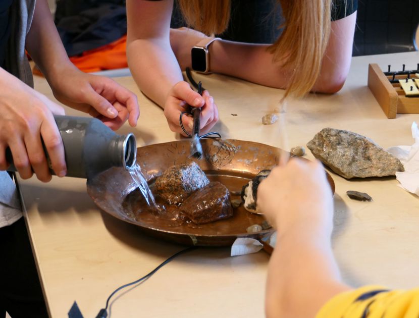 En gruppe mennesker laver lyd-eksperimenter med vand og sten i en skål. En person hælder vand fra en flaske over stenene, mens en anden holder et værktøj nær stenene.