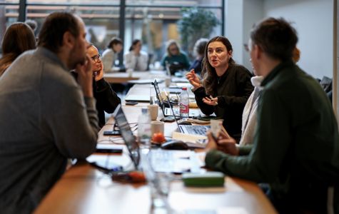 En gruppe mennesker sidder omkring et bord i et mødelokale. Der er bærbare computere, notesbøger og forskellige andre ting på bordet. I baggrunden er der flere mennesker og store vinduer, der lukker naturligt lys ind.