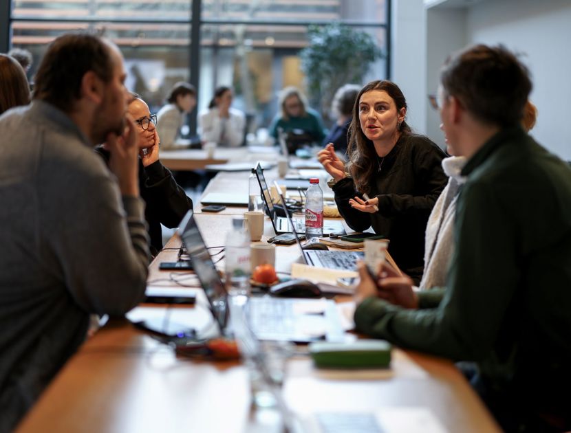 En gruppe mennesker sidder omkring et bord i et mødelokale. Der er bærbare computere, notesbøger og forskellige andre ting på bordet. I baggrunden er der flere mennesker og store vinduer, der lukker naturligt lys ind.