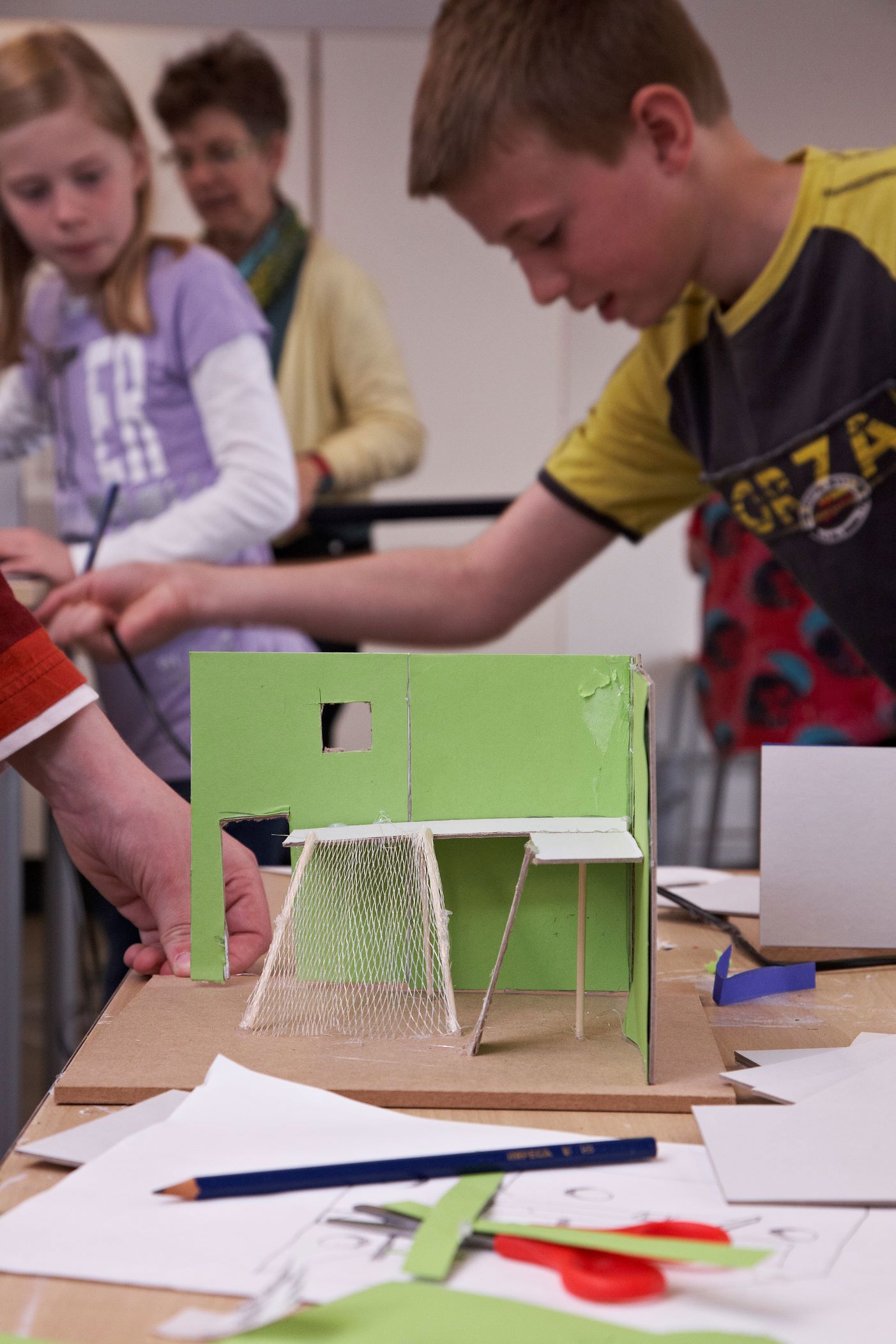 En gruppe børn arbejder på et projekt, der involverer en lille grøn modelstruktur. Modellen ser ud til at være lavet af pap og inkluderer elementer som vægge, et vindue og noget, der ligner net eller mesh-materiale. Der er forskellige papirer og værktøjer spredt på bordet omkring modellen.