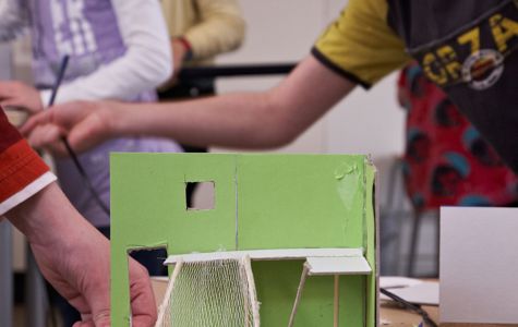 En gruppe børn arbejder på et projekt, der involverer en lille grøn modelstruktur. Modellen ser ud til at være lavet af pap og inkluderer elementer som vægge, et vindue og noget, der ligner net eller mesh-materiale. Der er forskellige papirer og værktøjer spredt på bordet omkring modellen.