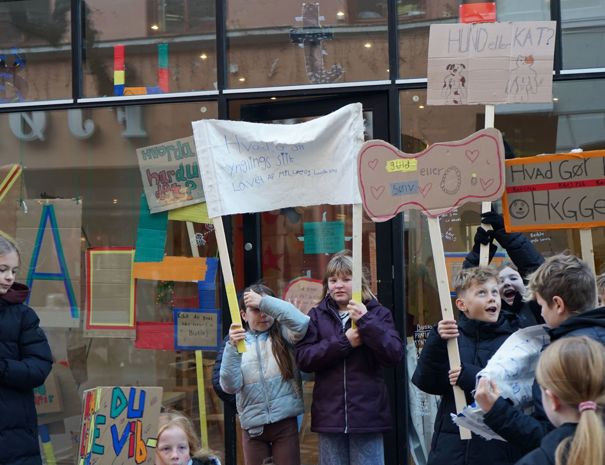 En gruppe børn holder forskellige skilte og bannere i en demonstration. Baggrunden inkluderer en butik med farverige dekorationer og bogstaver på vinduet.