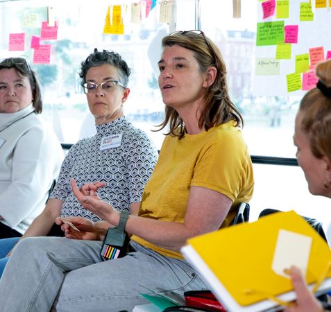 En gruppe mennesker sidder i et rum med store vinduer og deltager i en diskussion. Der er farverige post-it-noter på vinduerne bag dem, og en person holder en gul mappe og tager noter.