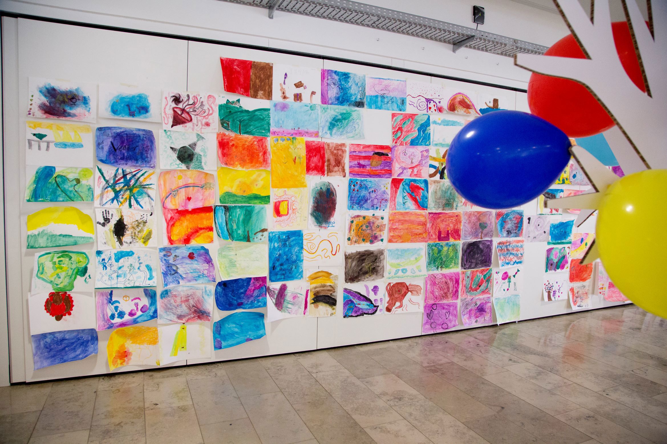 En væg dækket med mange farverige tegninger og malerier, hver på et separat ark papir. Kunstværkerne er arrangeret i et gittermønster. I forgrunden er der tre balloner: en blå, en gul og en rød. 