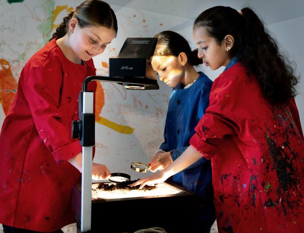 Tre personer iført røde og blå kitler står omkring en overheadprojektor. De er engageret i en aktivitet med projektoren. Baggrunden inkluderer en farverig væg med abstrakte mønstre eller tegninger.