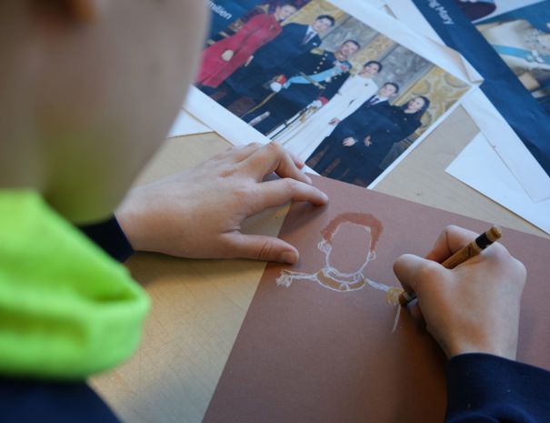 En person tegner på et brunt stykke papir med en hvid blyant eller farvekridt. Tegningen forestiller en person med fokus på hoved og skuldre. I baggrunden ligger der flere fotografier af personer i formelt tøj på bordet.