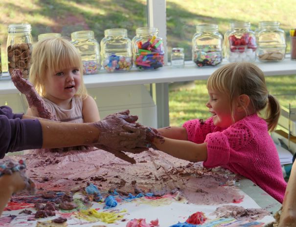 En gruppe børn, der leger med ler eller et andet formbart materiale ved et bord. Bordet er dækket med forskellige farver af materialet, og børnene bruger deres hænder til at forme det