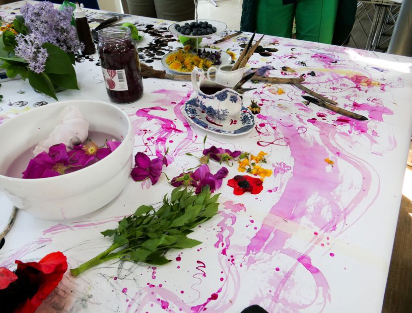 Et bord dækket med forskellige kunstmaterialer og forsyninger, herunder blomster, blade, en skål med vand og flydende kronblade, pensler, en tekop med underkop og spredte malerpletter i forskellige farver.