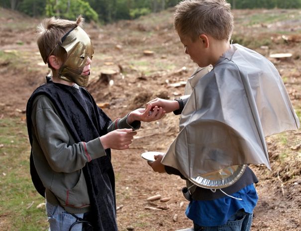 To børn i kostumer står i et skovområde. Den ene er klædt i sort med en guldmaske, og den anden bærer en sølvkappe med en tærteform på ryggen.