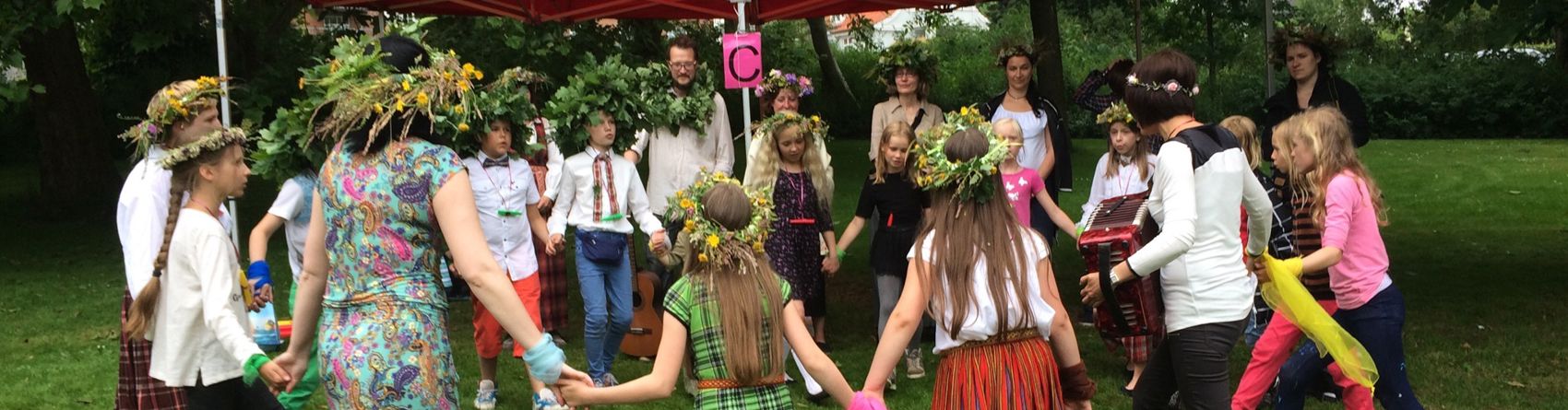 En gruppe mennesker, primært børn, deltager i en udendørs aktivitet under et rødt telt. Deltagerne holder hinanden i hænderne og danner en cirkel på græsset. Mange af dem bærer blomsterkranse og farverigt tøj.
