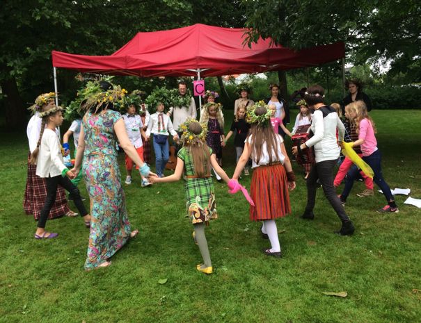 En gruppe mennesker, primært børn, deltager i en udendørs aktivitet under et rødt telt. Deltagerne holder hinanden i hænderne og danner en cirkel på græsset. Mange af dem bærer blomsterkranse og farverigt tøj.