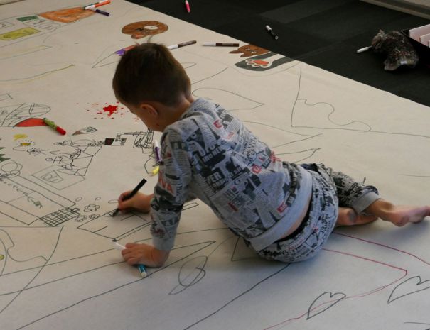 Et barn tegner på et stort ark papir, der er spredt ud på gulvet. Barnet er omgivet af forskellige farvede tuscher og farveblyanter, og der er flere tegninger på papiret, herunder hjerter, bygninger og andre former.