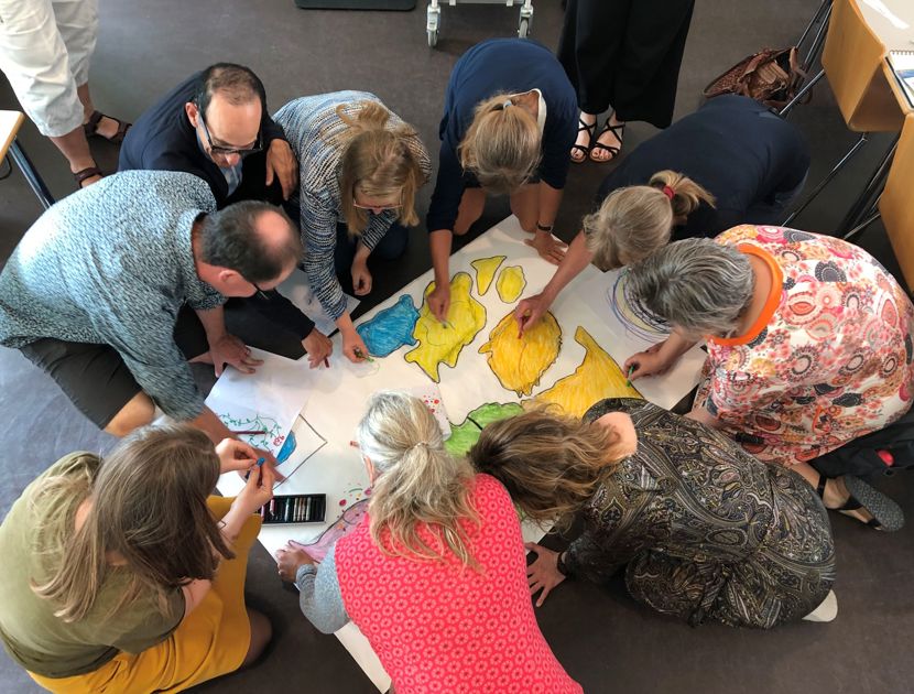 En gruppe mennesker er samlet omkring et stort stykke papir på gulvet med forskellige farvede sektioner. De er engageret i en aktivitet med papiret.