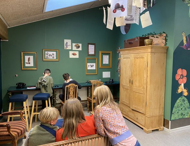 To børn sidder ved et skrivebord i et rum med grønne vægge og forskellige indrammede billeder. Bag dem er der et træskab og flere andre børn, der står eller sidder på møbler og observerer de to ved skrivebordet. Rummet har en hyggelig atmosfære med træmøbler og dekorative elementer som planter og kunstværker.