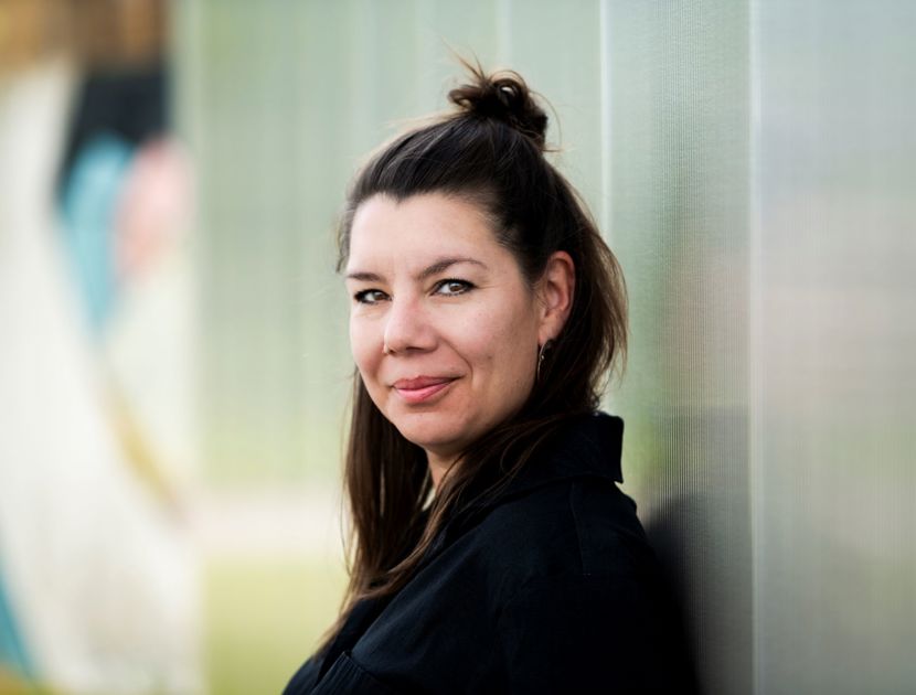 Camilla Høg står udendørs med håret sat op i en halv knold og læner sig mod en tekstureret væg. Baggrunden er udendørs med nogle slørede elementer.