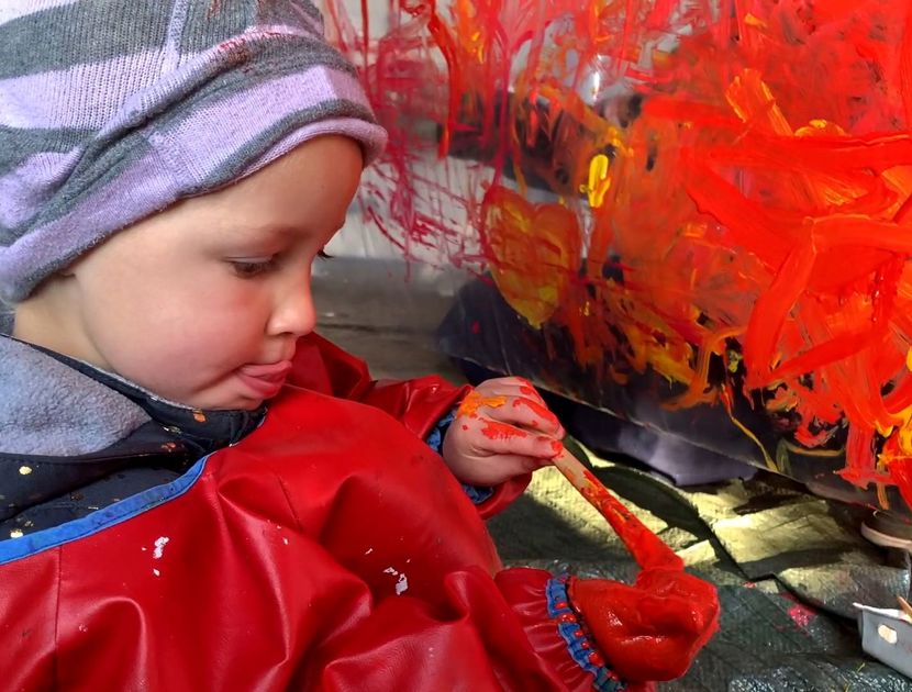 Et barn maler med rød og orange maling. Barnet er iført en rød malerkittel og en stribet hue. I baggrunden ses et lærred dækket af abstrakte røde og orange penselstrøg.