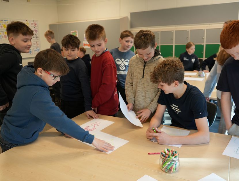 En gruppe elever i et klasseværelse. De er samlet omkring borde og engagerer sig i forskellige aktiviteter som tegning og diskussion. Nogle elever står op, mens andre sidder. Der er en beholder med farveblyanter på et af bordene.