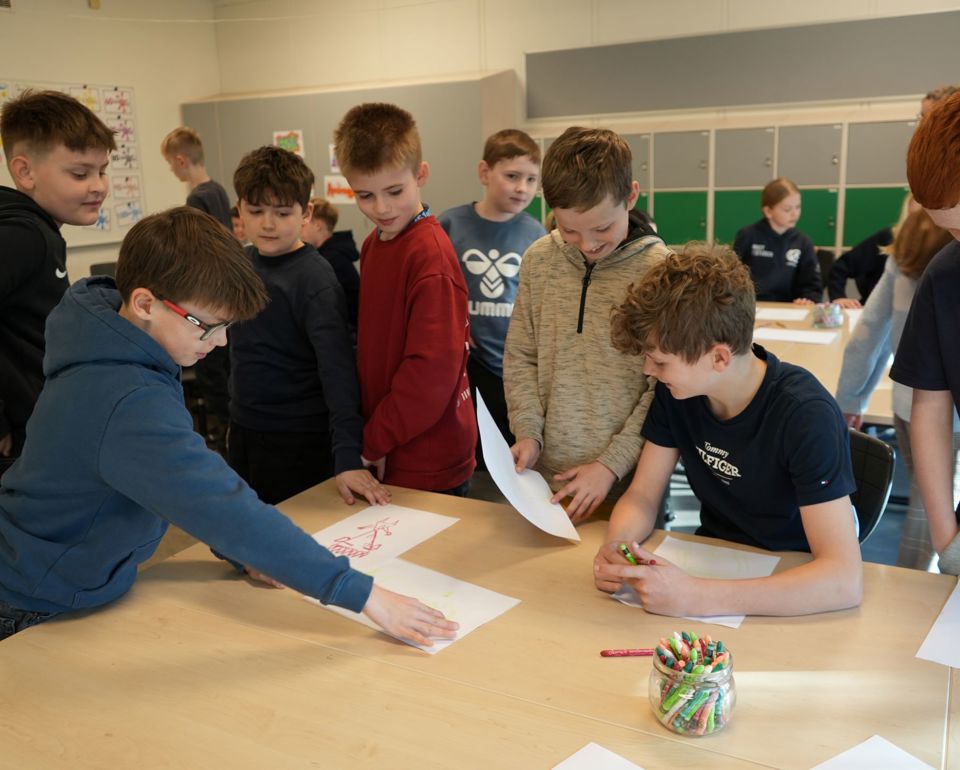 En gruppe elever i et klasseværelse. De er samlet omkring borde og engagerer sig i forskellige aktiviteter som tegning og diskussion. Nogle elever står op, mens andre sidder. Der er en beholder med farveblyanter på et af bordene.