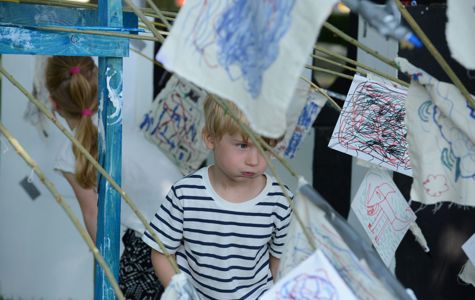 Et barn står blandt flere stykker stof eller papir med farverige tegninger og kruseduller, der hænger på snore. Barnet har en hvid og sort stribet trøje på.