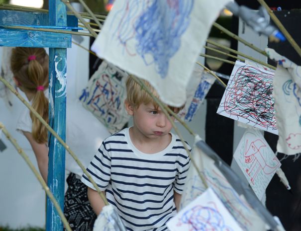 Et barn står blandt flere stykker stof eller papir med farverige tegninger og kruseduller, der hænger på snore. Barnet har en hvid og sort stribet trøje på.