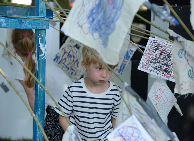 Et barn står blandt flere stykker stof eller papir med farverige tegninger og kruseduller, der hænger på snore. Barnet har en hvid og sort stribet trøje på.
