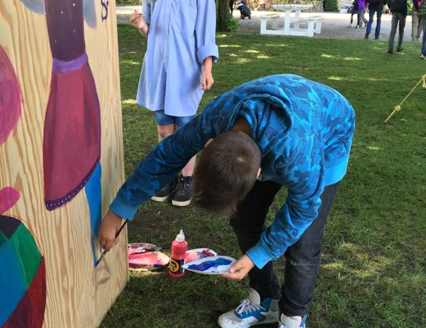To børn maler udendørs på en stor træplade med farverige kjolemotiver. Maleredskaber ligger på jorden omkring dem, og i baggrunden ses træer, bænke og en udendørs scene med mennesker.