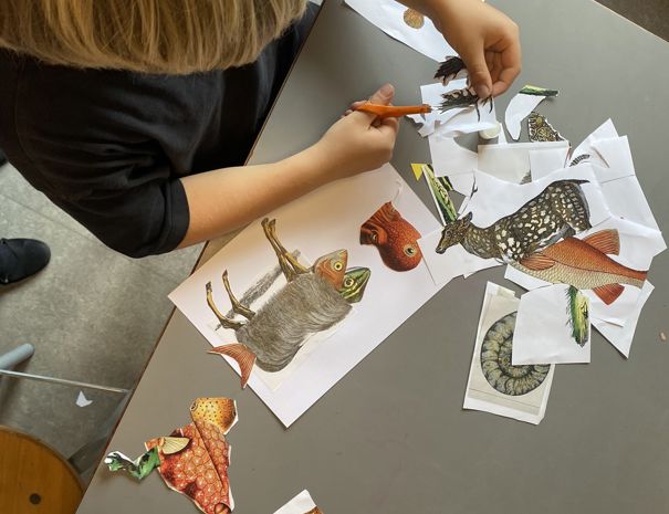 Et barn er engageret i en kunstaktivitet. Barnet klipper billeder af forskellige fisk og andre væsner ud fra trykte ark papir. Bordet er fyldt med udklippede stykker, saks og en limstift.