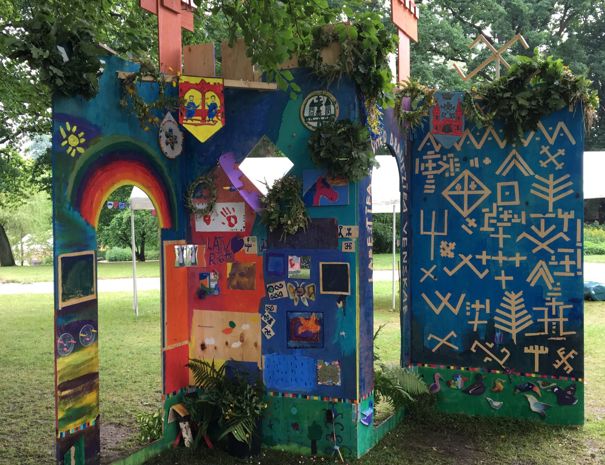 Farverige, kunstneriske byporte udendørs, der ligner et slot eller fort med to tårne. Strukturerne er dekoreret med forskellige symboler, mønstre og billeder, herunder en stor regnbue og intrikate geometriske mønstre.