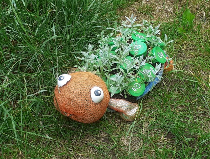 Et væsen lavet af affaldsmaterialer, med grønne blade som hår og små runde etiketter på kroppen, ligger på græsset ved siden af en brun netpose med to store øjne.