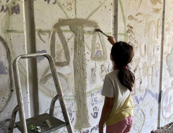 Et barn maler over graffiti på en væg med en pensel og mudder, og står ved siden af en stige.