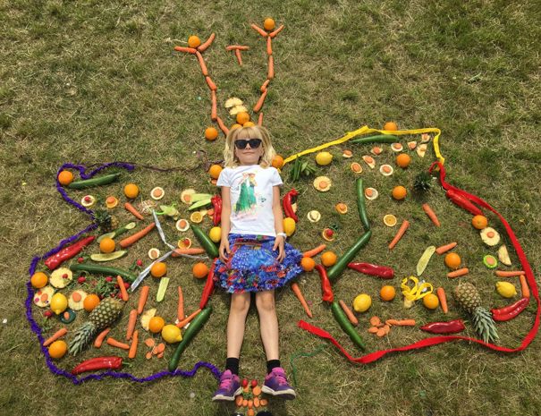 Et barn ligger på græsset med forskellige frugter og grøntsager arrangeret omkring sig i form af en sommerfugl.