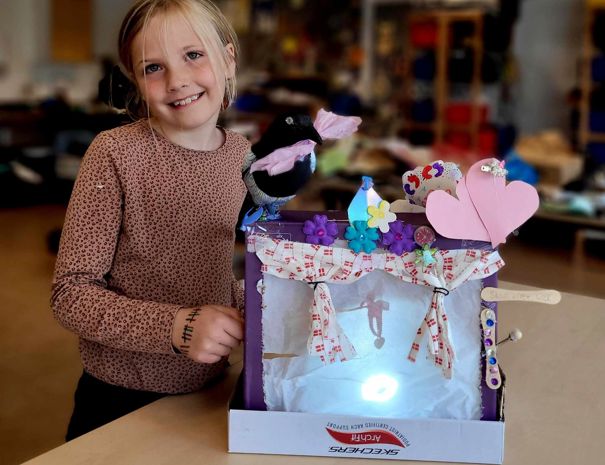 Et barn står i et klasseværelse og holder en dekoreret kasse. Kassen er pyntet med forskellige materialer som blomster, hjerter og bånd. Inde i kassen er der en lille figur, der hænger i midten med lys, der skinner på den.