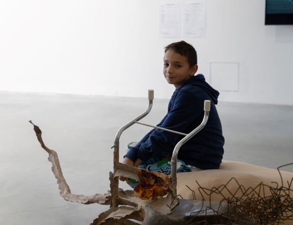 Et barn sidder på en abstrakt skulptur lavet af metal og andre materialer. Skulpturen inkluderer snoede metalstænger og en struktur, der ligner styr.